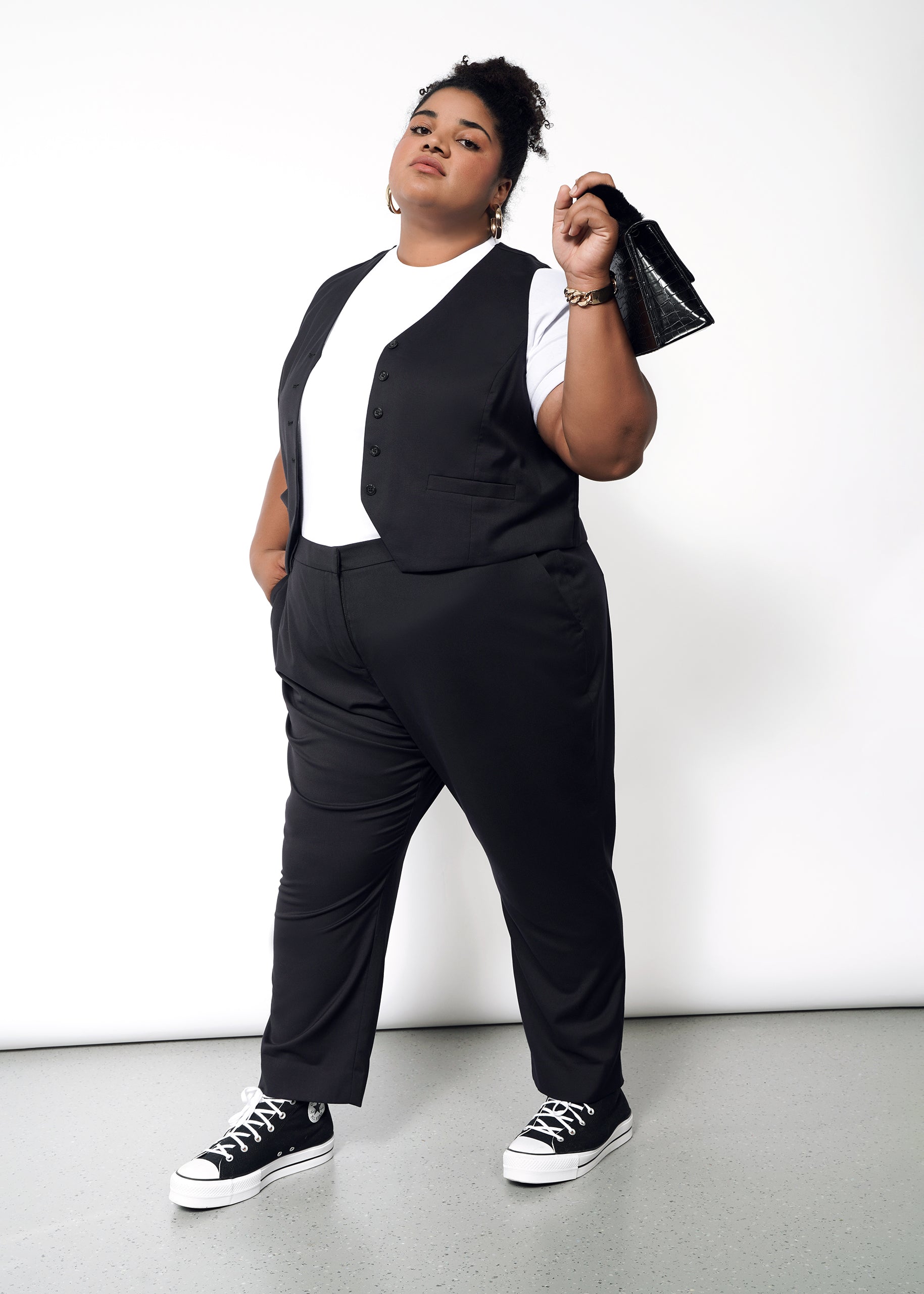 Confident individual against a white backdrop in The Empower Vest over a white t-shirt, paired with black pants and sneakers. A small black handbag is held effortlessly with one hand in their pocket, showcasing style and comfort.