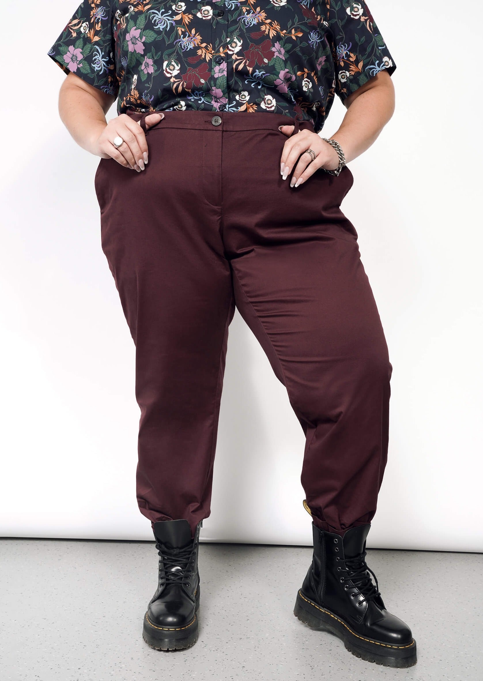 A person in a floral shirt and The Essential Trouser with a burgundy, tapered leg stands with hands in pockets. They wear black boots on a grey speckled floor against a plain white background.