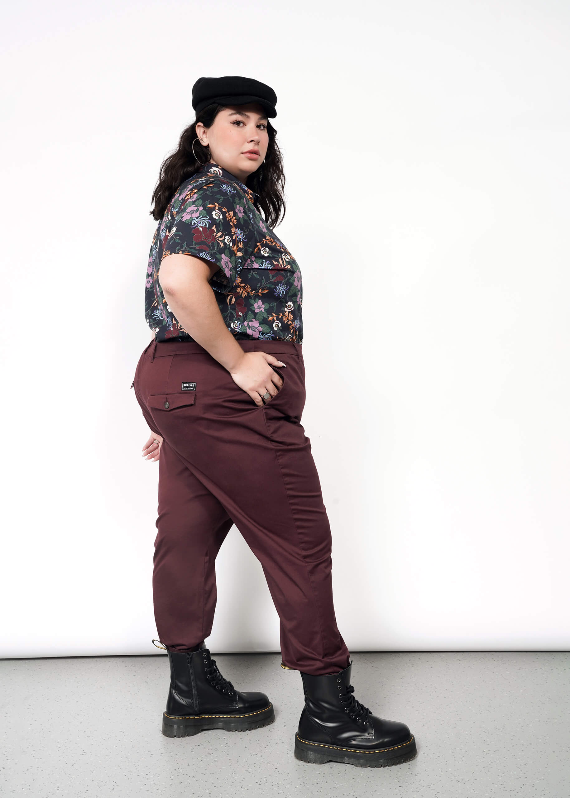 A person stands confidently against a white background, hands in pockets, slightly turned to the side. They wear a black cap, floral shirt, The Essential Trouser in maroon with a tapered leg, and black boots.