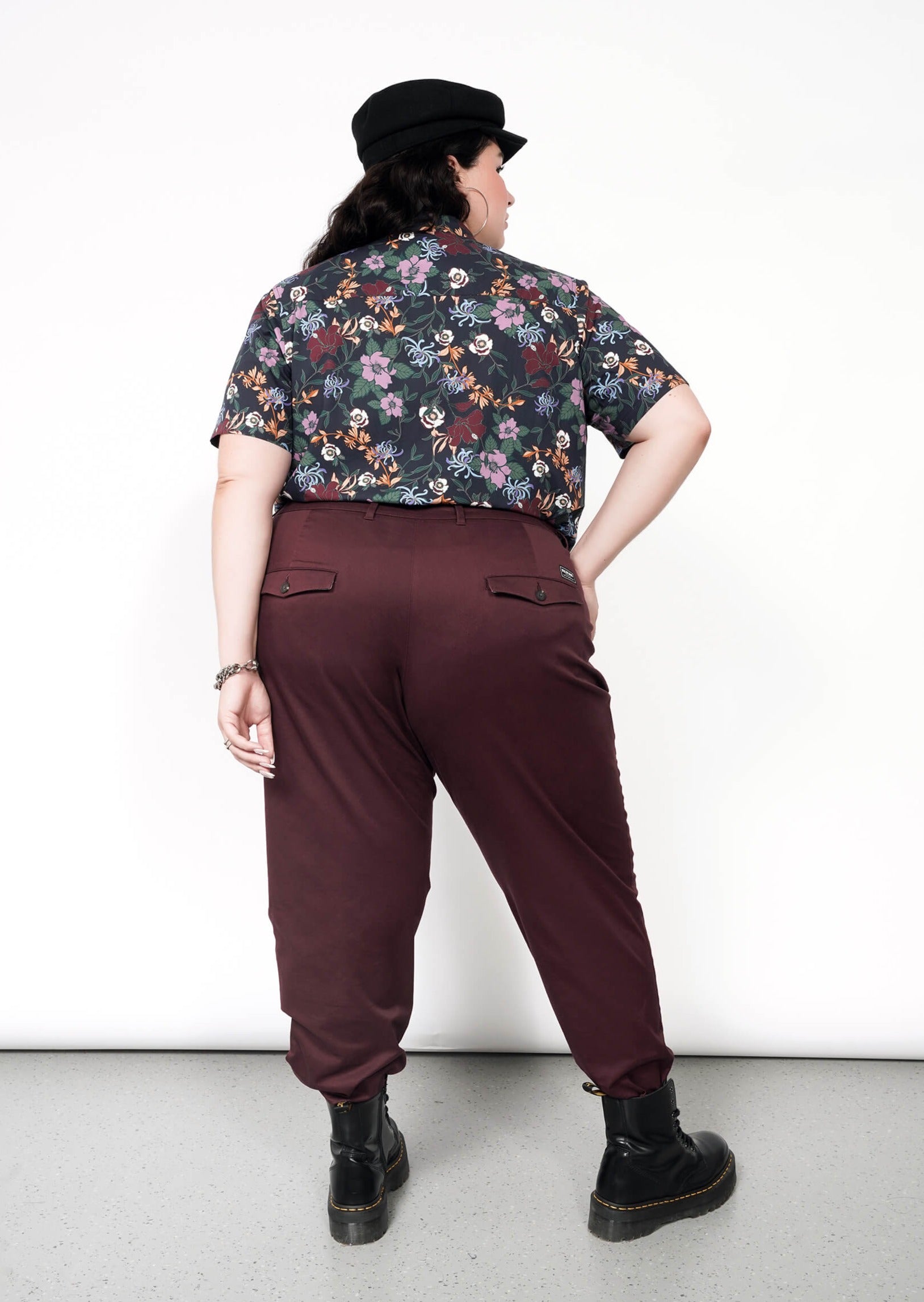 Standing against a plain white background, a person faces away wearing The Essential Trouser in classic maroon with a tapered leg, paired with a floral shirt and black cap. Black boots complete the ensemble as they pose with one hand on their hip.