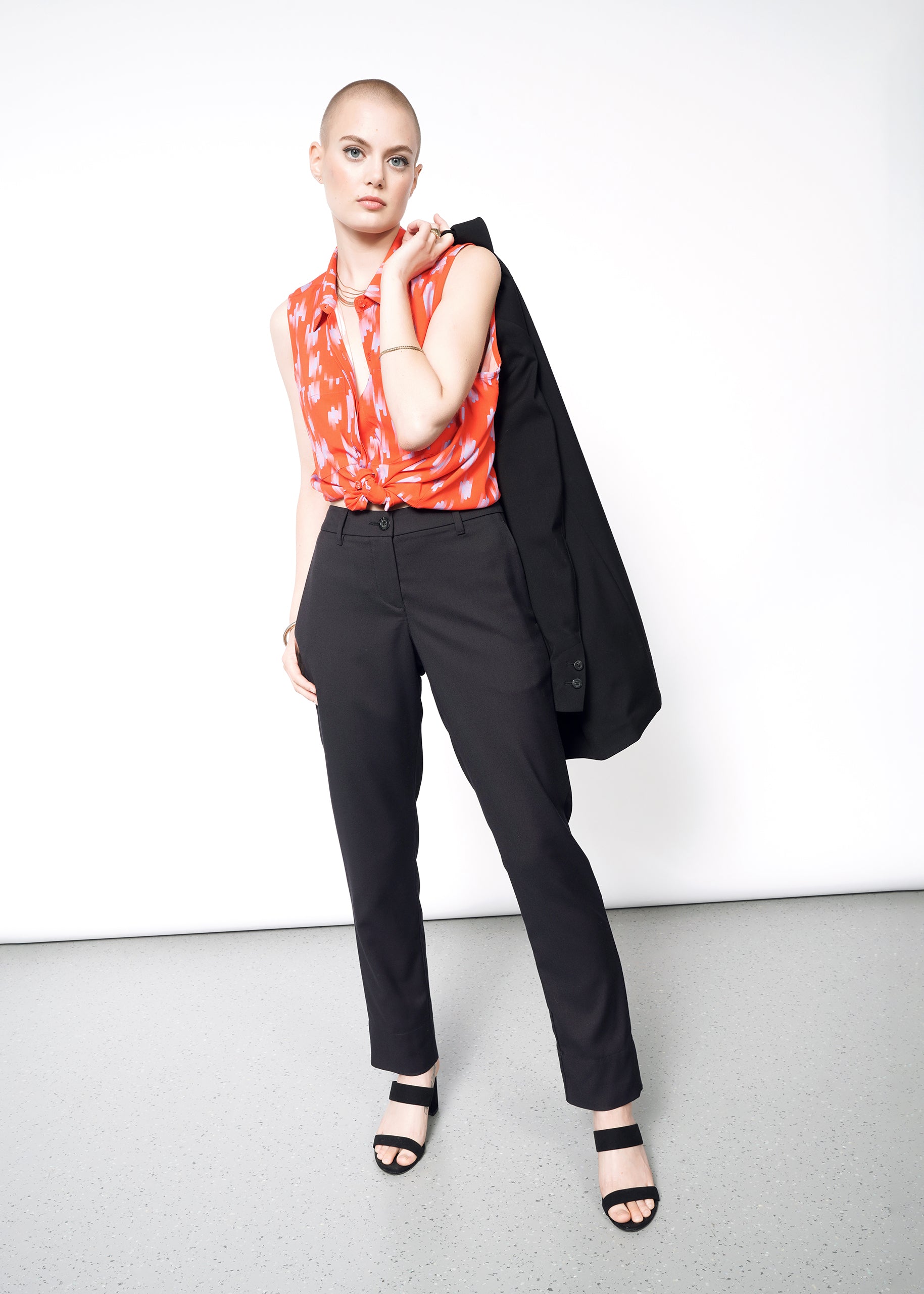 A bald person poses confidently in The Empower Trouser, a red patterned top, and sandals, holding a black jacket over one shoulder against a plain white wall and gray floor backdrop.