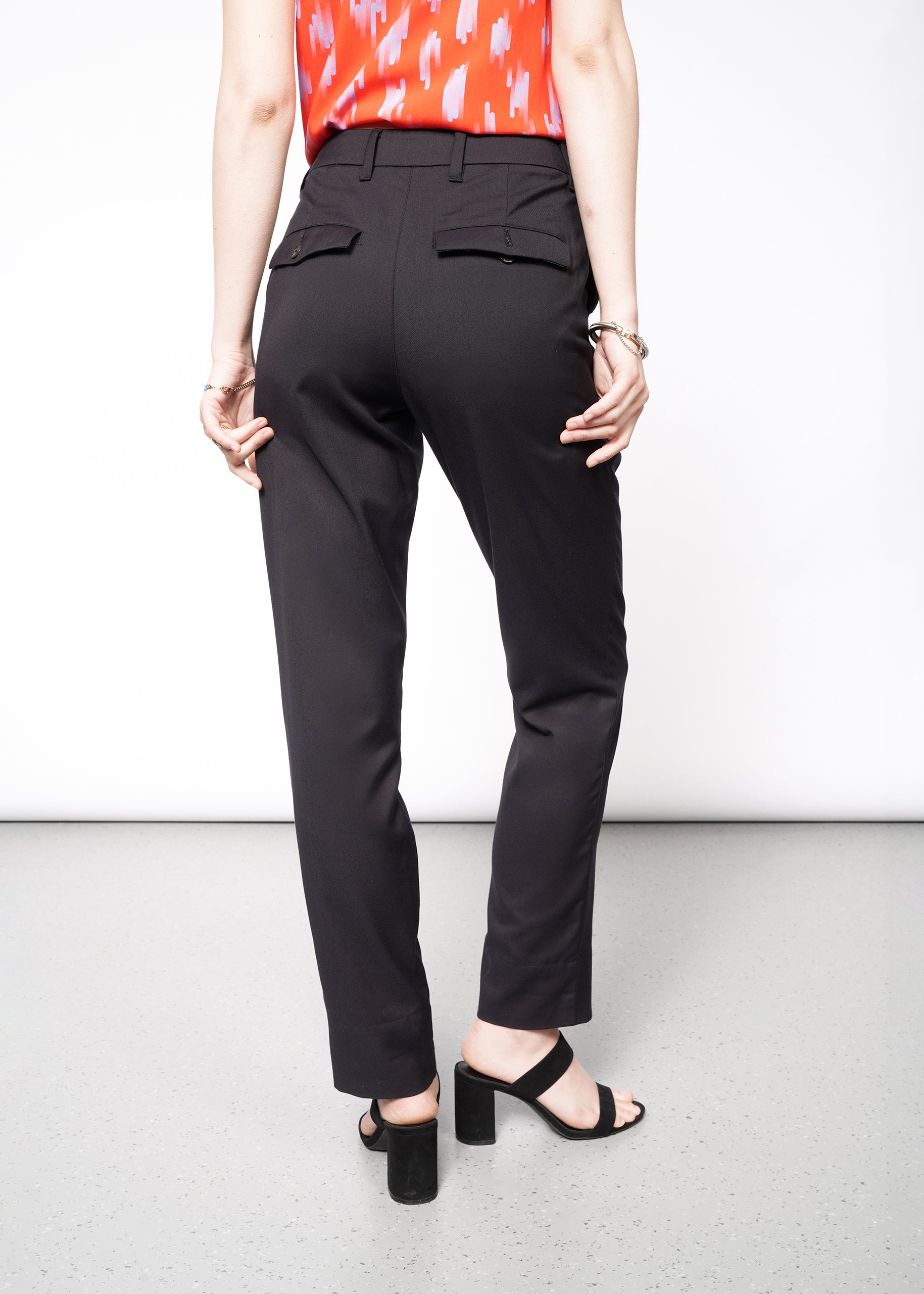 Dressed in The Empower Trouser, sleek black mid-rise pants with a tapered leg and rear pockets, the person stands on a light-colored floor wearing a red patterned top and black heels. Their hands rest relaxed by their sides.