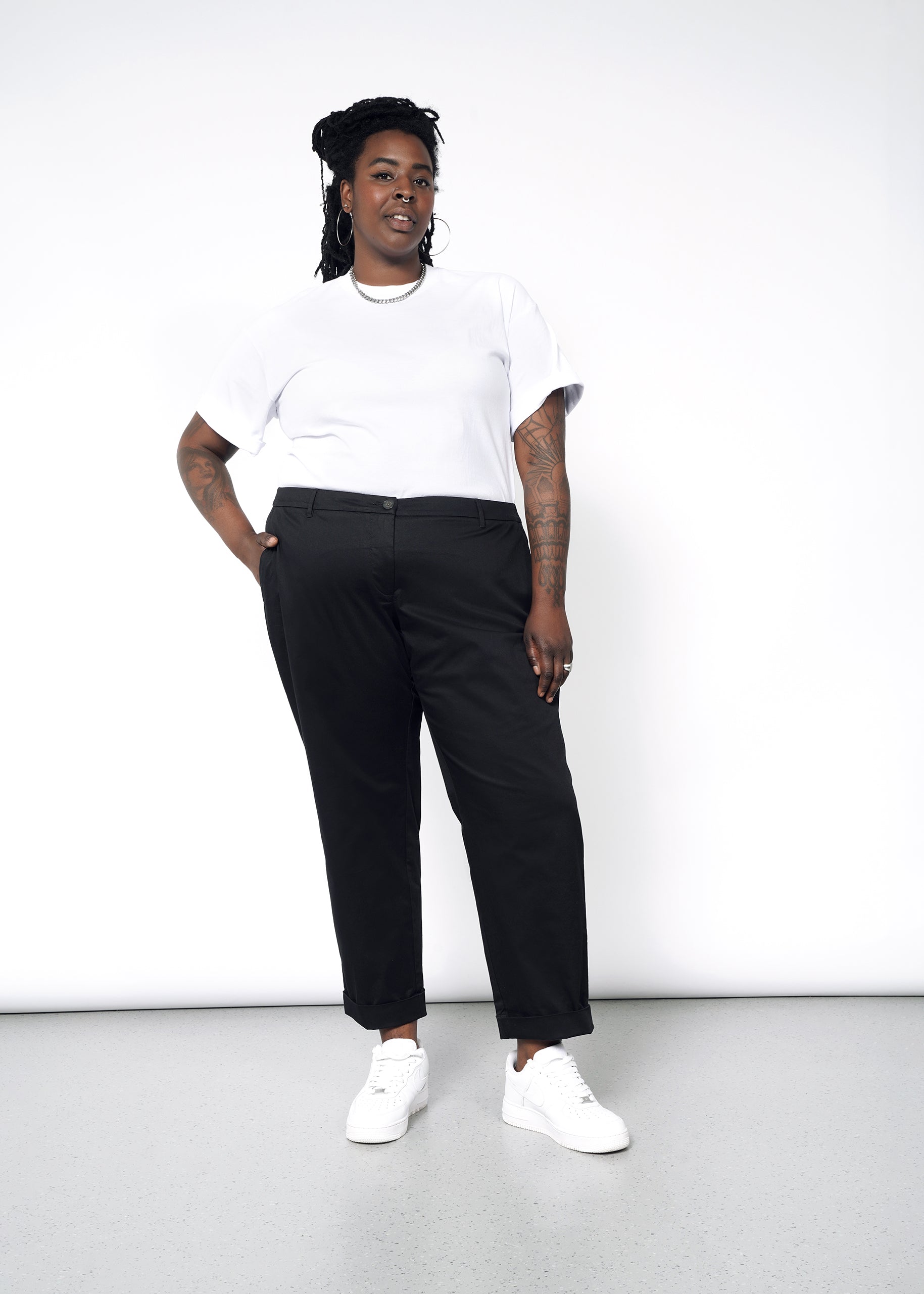 A person with braided hair stands confidently against a white background, wearing a white t-shirt, The Essential Trouser with a relaxed mid-rise fit, and white sneakers. Their hand is in their pocket, and they are smiling.