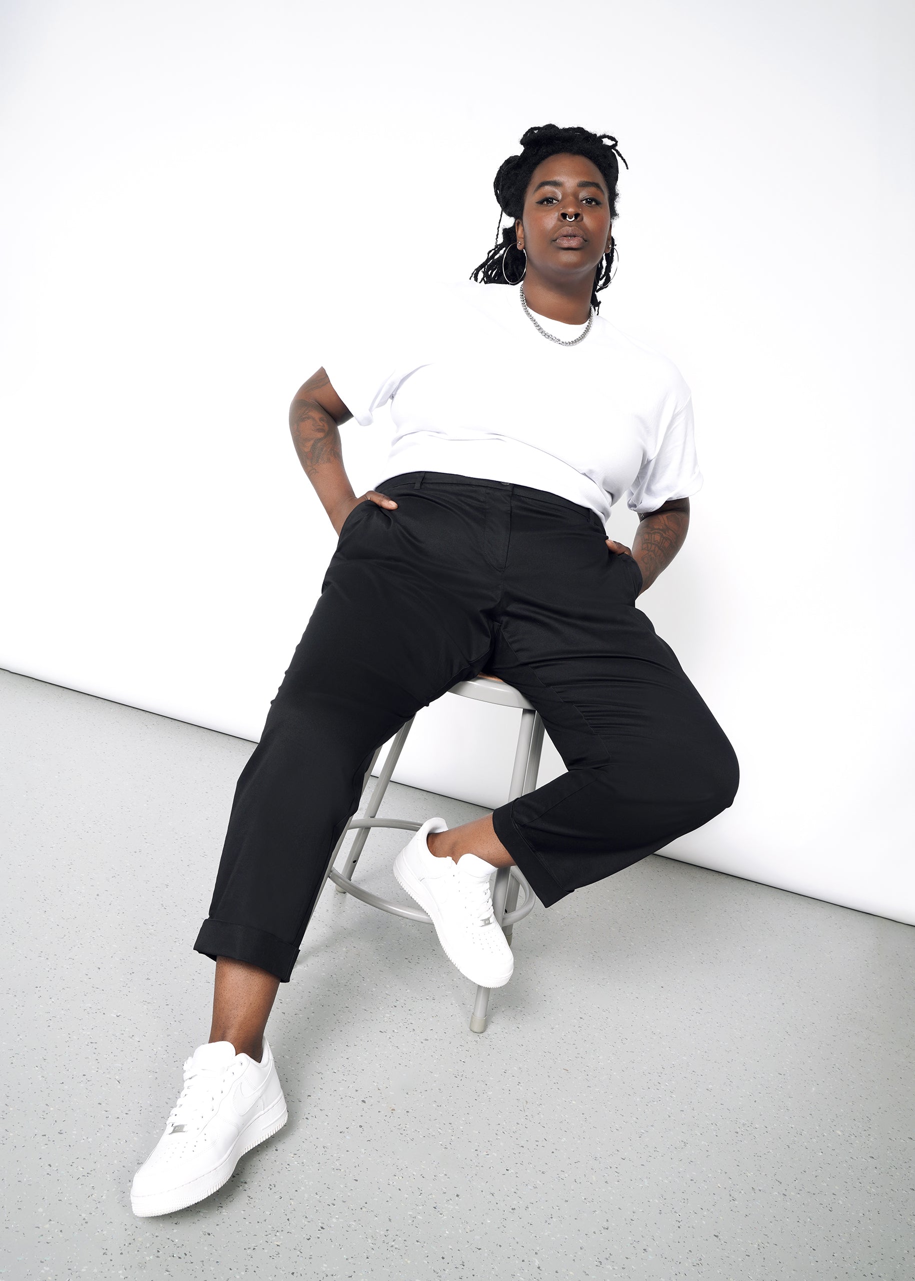 A person sits confidently on a stool against a white backdrop, dressed in a white t-shirt and The Essential Trouser, paired with white sneakers. With one leg extended and hands in pockets, they look directly at the camera.